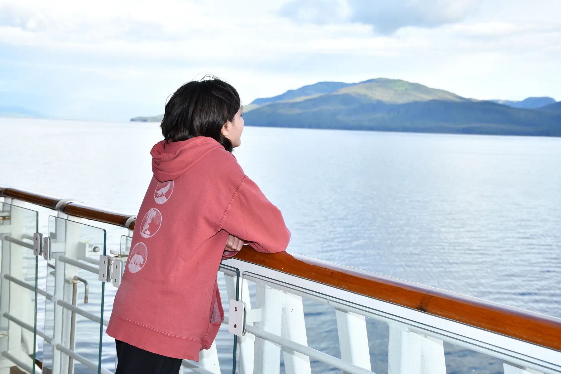 Ronan leaning on a ship railing in a red hoodie, looking out at mountains in Alaska